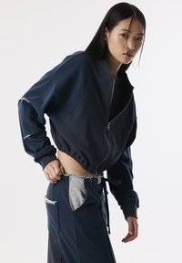Donkerblauwe cropped jack met een ritssluiting aan de voorkant, geribbelde textuur en brede mouwen. Gecombineerd met bijpassende broek met zilveren zakaccenten.