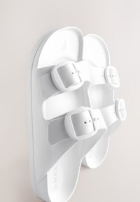 Sandales blanches avec deux boucles réglables, en matériau synthétique texturé ; semelle plate ; design simple avec orteil et talon ouverts.