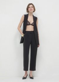 Schwarze maßgeschneiderte Weste ohne Ärmel über einem Spitzen-BH, kombiniert mit hochgeschnittenen, schmal zulaufenden Hosen und spitzen, schwarzen Absätzen. Akzente in Gold an den Armreifen.