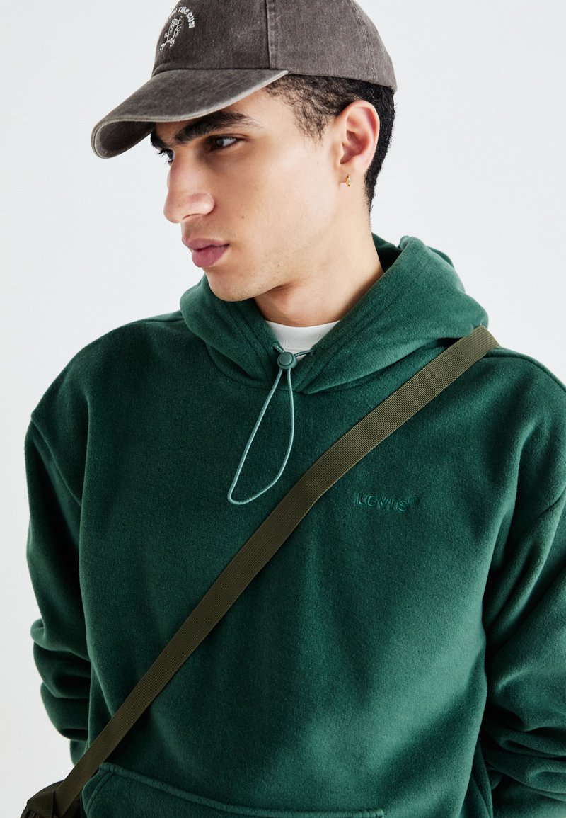 Dunkelgrüner Hoodie mit Kordelzug, aus weichem Material und mit gesticktem Logo. Kombiniert mit einer grauen Baseballkappe und einem braunen Riemen.
