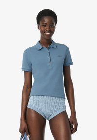 Vaaleansininen polo-paita, jossa on kaulus, neljän napin napitus ja hillitty logo. Yhdistetty kuviollisiin vaaleansinisiin shortseihin, joissa on paksun neuleen muotoinen kuvio.