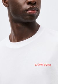 Κοντινό πλάνο ενός ατόμου που φοράει λευκό T-shirt με κόκκινο κείμενο "BJÖRN BORG" στο αριστερό στήθος, κοιτάζοντας ελαφρώς προς το πλάι.