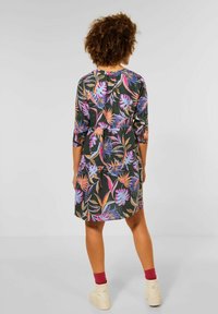Kwiatowa sukienka z ciemnym tłem i kolorowymi wzorami tropikalnych liści, z rękawami 3/4 i luźnym krojem. Noszona z beżowymi butami.