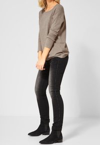 Pull marron tricoté avec un large col, associé à un jean foncé avec des détails de couture sur le côté, et des bottines noires.