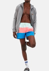 Chaqueta gris con cremallera y franjas laterales, llevada sobre bañador multicolor (rosa, azul, menta). Calcetines tobilleros blancos con rayas azules, chanclas negras.