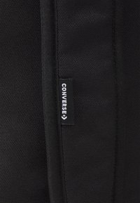 Detail eines schwarzen Stoff-Rucksacks mit strukturiertem Oberfläche und einem kleinen, weißen Converse-Logosticker, der in die Kante genäht ist.