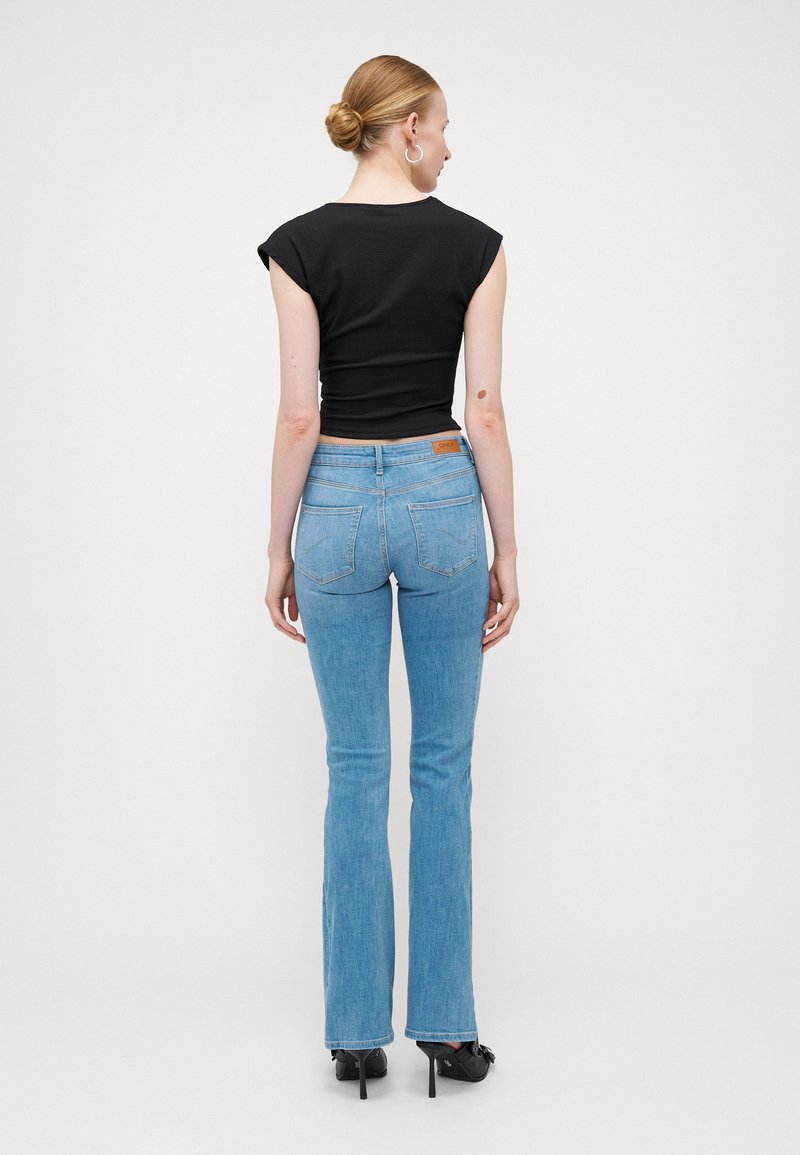 Hoch taillierte hellblaue Jeans mit weitem Bein und dezenten Nähten. Kombiniert mit einem figurbetonten schwarzen Oberteil und Absatzschuhen.