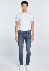 Młody mężczyzna stoi prosto, ubrany w dopasowaną białą koszulkę, niebieskie dżinsy i białe trampki na jednolitym jasnym tle.