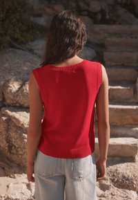 Žena s dlhými tmavými vlasmi, oblečená v červenom bezrukávovom svetri a svetlomodrých džínsach, stojaca vonku pri kamenných schodoch a skalách.