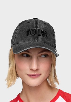 Joven mujer con cabello rubio corto que lleva una gorra de béisbol negra descolorida con bordado "TOUS" y un detalle de cadena plateada en la visera.