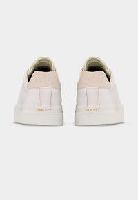 Ζευγάρι λευκά αθλητικά παπούτσια χαμηλού ύψους με μπεζ σουέντ ταμπόν στη φτέρνα και υφή στη σόλα, με θέα από πίσω σε λευκό φόντο.