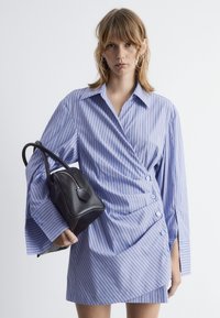 Gestreiftes Hemdkleid in Blau und Weiß mit übergroßen Ärmeln, Knopfdetails und Wickeldesign. Kombiniert mit einer kleinen schwarzen Lederhandtasche.