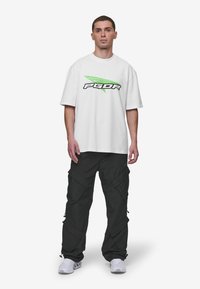 Bijela oversized majica s crnim i zelenim "PGDR" logotipom, uparena s crnim cargo hlačama s džepovima i patentnim zatvaračima, nošena uz bijele tenisice.
