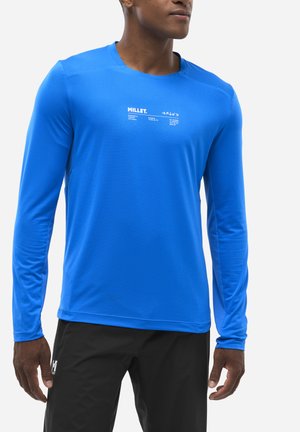 Homme portant une chemise Millet bleu vif à manches longues et un pantalon de sport noir, debout les bras détendus le long du corps.