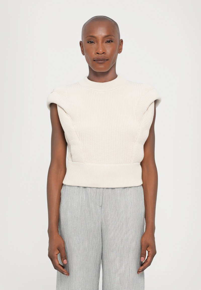 Knit cream-colored sleeveless sweater with ribbed texture and structured shoulder design, paired with light-striped wide-leg pants.
