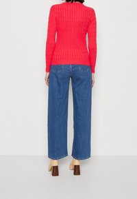 Woman wearing a red cable knit sweater, blue wide-leg jeans, and beige heeled ankle boots, standing against a plain white background.