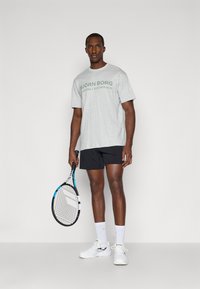 Sivá bavlnená tričko s zeleným potlačou, čierne šortky, biele športové topánky a drží tenisovú raketu s modrými akcentmi. Minimálny pozadie.