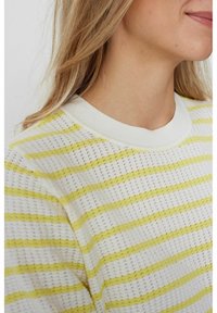 Částečný pohled na osobu oblečenou v bílém pleteném svetr s žlutými vodorovnými pruhy a kulatým výstřihem.