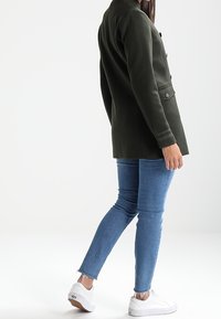 Tmavě zelený, vykrojený blejzr s jednořadým zapínáním, dvěma předními kapsami, v kombinaci s přiléhavými modrými džíny a bílými teniskami.