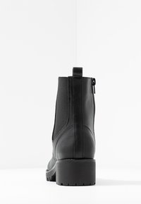 Bottine noire en cuir avec des panneaux latéraux élastiques, une fermeture éclair et un talon épais. La vue arrière met en évidence des surfaces lisses et texturées.