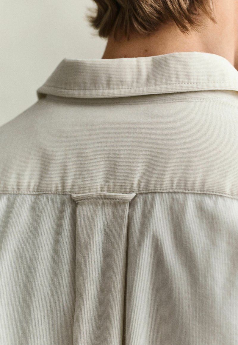 Back view of a person wearing a beige corduroy shirt with a collar and a centrally stitched back yoke.