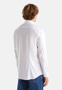 Camicia bianca a maniche lunghe realizzata in tessuto morbido, con colletto alla francese, polsini con bottone e orlo arrotondato. La vista posteriore mostra un design minimalista.