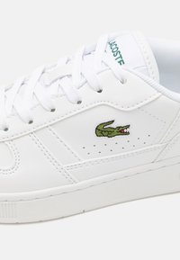 Hvit lær sneakers med teksturert overflate, med en grønn krokodille-logo og hvite snørebånd, designet med perforerte detaljer på sidene.