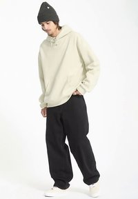 Sweat à capuche beige avec cordon de serrage, poche kangourou, et pantalon ample noir. Bonnet en tricot gris et baskets blanches complètent la tenue.
