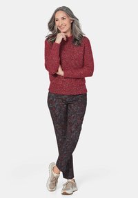 Rode gestructureerde gebreide trui met een geribbelde zoom, gecombineerd met een broek met een donker bloemenpatroon. Draag het met beige sneakers terwijl het model met een glimlach poseert.