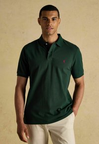 Sötétzöld, rövid ujjú pólóing, gallérral, a mellkasán egy kis rózsaszín logóval. Simavászon, klasszikus fazon.