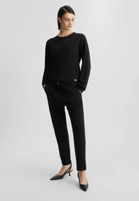 Zwarte sweatshirt met studded halslijn, gecombineerd met zwarte tapered broeken met gouden knoopaccenten en zwarte slingback hakken.