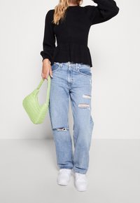 Pull noir côtelé avec ourlet peplum, associé à un jean bleu clair usé et un sac vert tressé. Des baskets blanches complètent le look.
