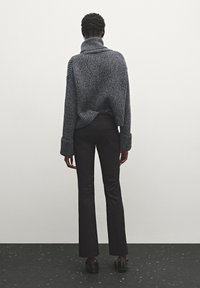 Pull tricot gris épais avec un col montant, une coupe ample et de larges poignets, associé à un pantalon noir évasé, vu de dos.