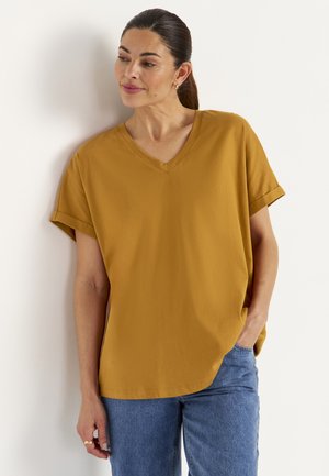 Vrouw met donker haar in een paardenstaart, gekleed in een mosterdgele V-hals T-shirt en blauwe jeans, staand tegen een witte muur met één hand in haar zak.