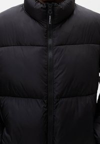 Veste matelassée noire avec un col montant, des sections horizontales cousues et une fermeture éclair complète à l'avant. Tissu lisse et brillant sans ornements visibles.