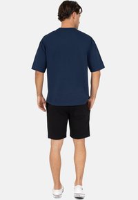 Heren t-shirt met korte mouwen in marineblauw en een ronde halslijn, gecombineerd met zwarte shorts en witte sneakers, gezien van achteren.