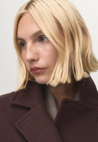 Primo piano di una donna con capelli biondo chiaro e ondulati e pelle chiara, che indossa un cappotto marrone scuro con colletto alto. Ha occhi azzurri chiari e un'espressione neutra.