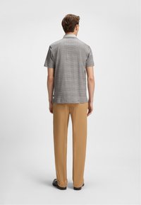 Hombre vestido con una camisa de cuadros gris de manga corta, pantalones beige y zapatos negros, de pie de espaldas contra un fondo ligero y liso.