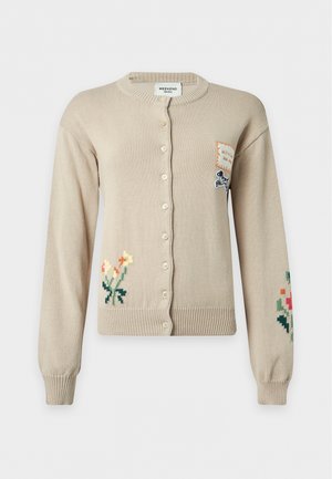 Frau steht mit den Händen hinter dem Rücken, trägt eine beige geknöpfte Strickjacke mit Blumenstickerei und weite weiße Hosen mit schwarzen spitzen Schuhen.