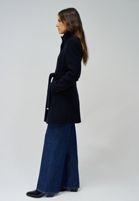 Manteau en laine bleu marine avec un col montant et une taille ceinturée, associé à un jean en denim coupe large et des bottines noires. Texture douce et coupe ajustée.