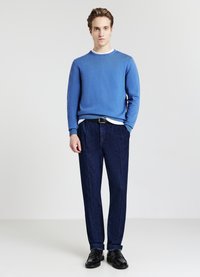 Junger Mann, der mit einer Hand in der Tasche steht, trägt einen blauen Pullover, ein weißes Hemd, dunkelblaue Hosen, einen schwarzen Gürtel und schwarze Schuhe vor einem einfarbigen Hintergrund.