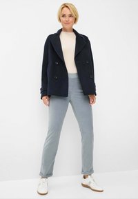 Marineblauwe dubbelgeknoopte zeemanjas, geribbelde lichte trui, lichtblauwe broek en witte sneakers met grijze accenten, tegen een effen witte achtergrond.