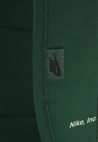 Grön Nike-textil med en strukturerad yta, sömdetaljer och en enda logotags med svarta bokstäver mot en mörkare grön bakgrund.