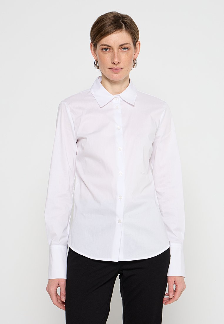 Weißes Langarmhemd mit Knopfleiste, klassischem Kragen und glattem Baumwollstoff. Getragen mit schwarzen Hosen.