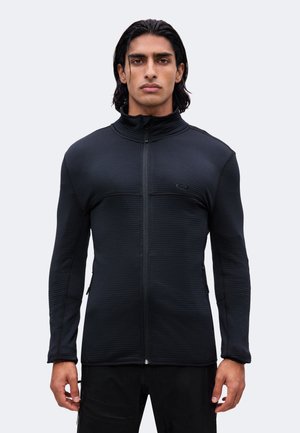 Hombre con cabello oscuro hasta los hombros que lleva una chaqueta negra ajustada con cremallera y pantalones negros, de pie frente a un fondo claro y liso.