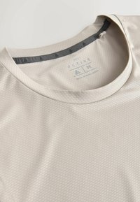 T-shirt de sport beige clair texturé avec encolure ronde et étiquette intérieure imprimée indiquant la taille M et "Fabriqué au Bangladesh."