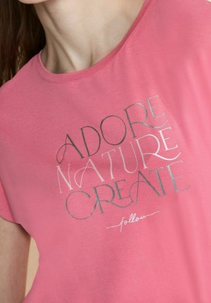 Femme portant un t-shirt rose avec le texte « ADORE NATURE CREATE » et « follow » en écriture plus petite en dessous.