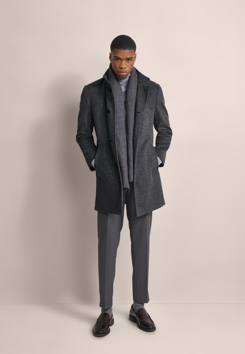 Cappotto di lana grigio con colletto a revers, abbinato a un dolcevita grigio chiaro e pantaloni coordinati. Scarpe lucide scure completano il look.