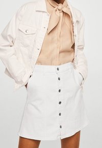 Jupe en denim blanc de forme évasée, avec une fermeture à boutons à l'avant et des poches, associée à un blouse beige clair et une veste pâle.