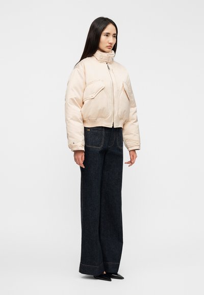 Orezaná svetlo béžová prešívaná bunda s vysokým golierom a dvoma prednými vreckami, kombinovaná s tmavými širokými džínsami a čiernymi topánkami so špicatou špičkou.